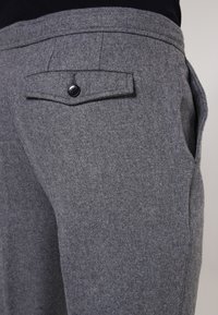 Close-up of gray wool trousers pocket with flap secured by a single black button, worn with a dark top.