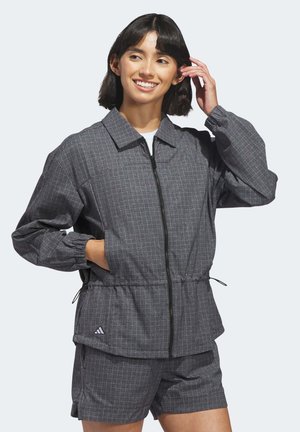 Smiling woman with short dark hair wearing a gray checkered zip-up jacket and matching shorts, hand in pocket and touching hair.