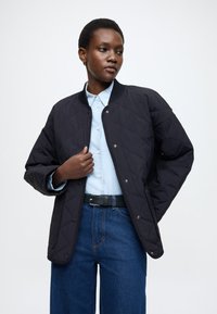 Crna prošivena bomber jakna s kopčama na pritisak, nošena preko svijetloplave košulje, uparen s tamnoplavim trapericama. Glatka tekstura, opušteni kroj.