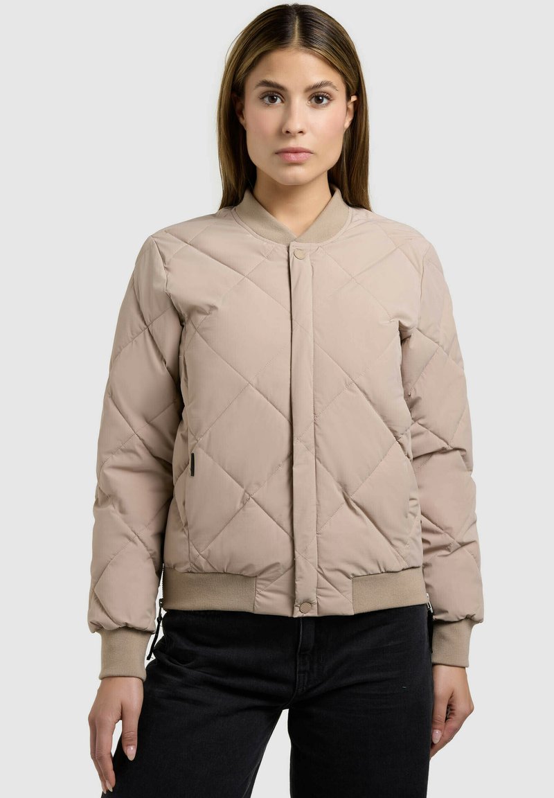 Femme portant une veste bomber matelassée beige avec des manches et une ceinture côtelées, associée à un pantalon noir, debout devant un fond uni.