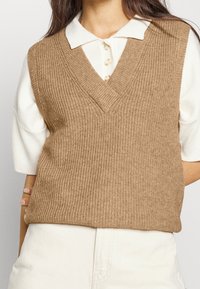 Bruine geribbelde gebreide mouwloze trui met een V-hals, gedragen over een wit overhemd met kraag, met een textuurrijke, gezellige uitstraling.