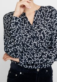 Žena v tmavě modré blůze s dlouhým rukávem a bílým květinovým vzorem, v černých manšestrových kalhotách, s překříženýma rukama a jednou rukou, která se dotýká její brady.