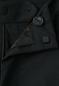 Pantalones negros con cierre de cremallera, botón de sujeción y detalle de lazo para cinturón de tela, que presentan un material suave y texturizado.
