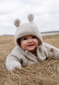 Hellbeige Fleecejacke mit Kapuze und Bärenohren. Das Baby ruht auf Stroh, lächelt, mit weicher Textur und einfachem Design.