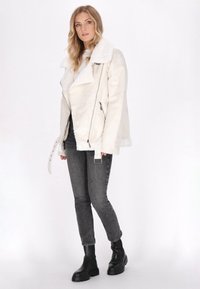 Kremowa kurtka z faux shearling o szerokim kołnierzu, zamek błyskawiczny i regulowany pasek. Noszona z szarymi dżinsami i czarnymi, masywnymi butami.