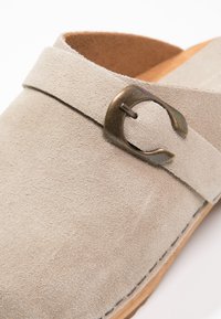 Beige Wildlederschuh zum Hineinschlüpfen mit runder Form, Holzsohle und einem bronzefarbenen Schnallenakzent am Riemen. Strukturierte Oberfläche mit Nahtdetails.