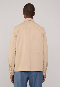 Beige Baumwolljacke mit spitzem Kragen, geknöpften Manschetten und Nahtdetails. Schlichtes Design, Rückansicht, getragen über blauen strukturierten Hosen.