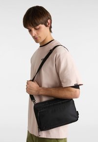 Bolso cruzado de nailon negro con dos compartimentos con cremalleras, diseño plano, correa ajustable, sostenido por una persona que lleva una camisa de color rosa claro.