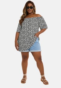 Off-shoulder topp i svart och vitt blommigt mönster, ihop med ljusblå denimshorts. Modellen bär flata sandaler och stora solglasögon.