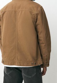 Giacca in tela marrone con un design strutturato, dotata di colletto, bottoni a pressione all'orlo e una texture liscia. Indossata su jeans scuri.