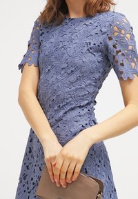 Vestido de encaje en azul claro con patrones de flores recortados, manga corta y corsé ajustado. La modelo sostiene un bolso de mano beige.