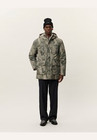 Abrigo acolchado con estampado de camuflaje en tonos tierra, con capucha, dos bolsillos delanteros y cierre de cremallera. Se lleva con pantalones oscuros y un gorro.