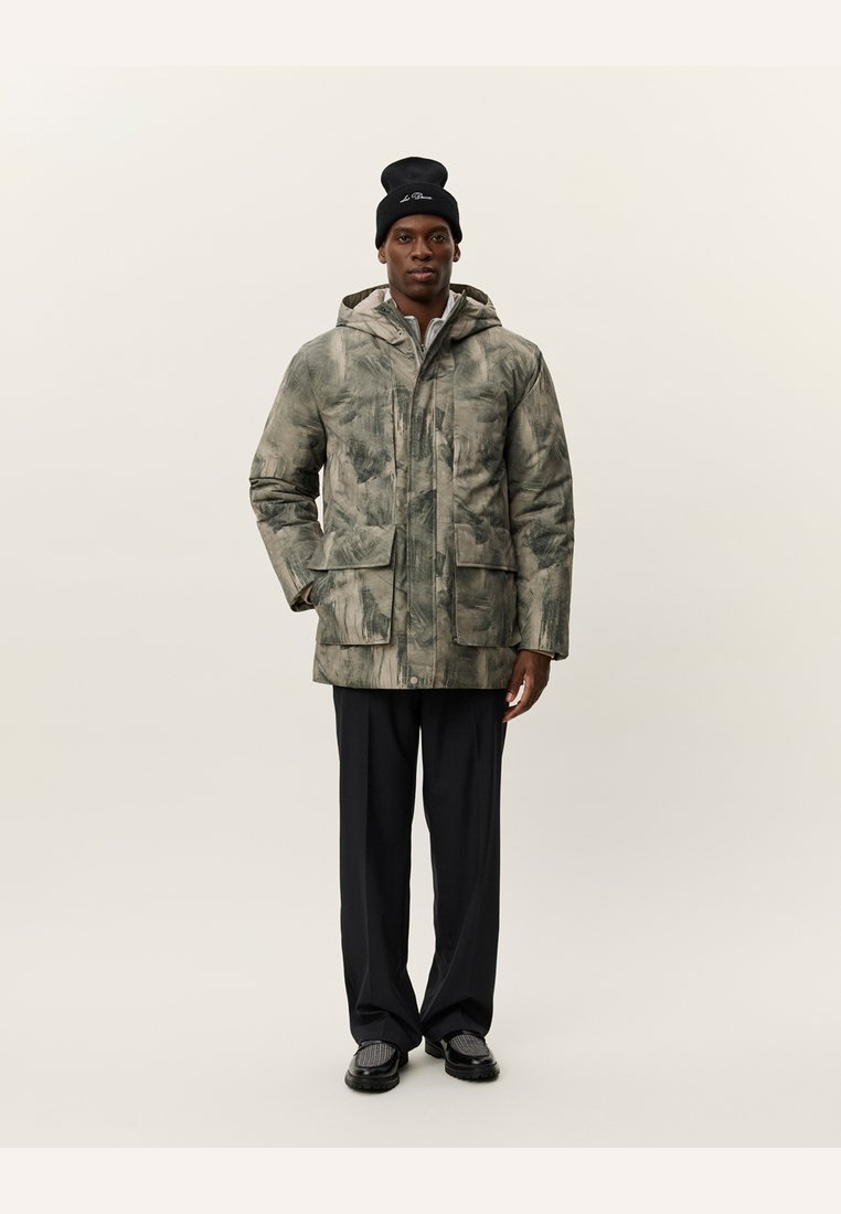 Abrigo acolchado con estampado de camuflaje en tonos tierra, con capucha, dos bolsillos delanteros y cierre de cremallera. Se lleva con pantalones oscuros y un gorro.
