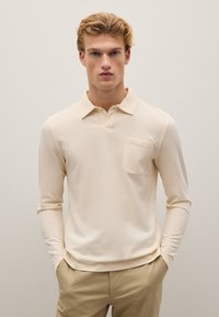 Beige Langarm-Polohemd mit Kragen und einer Brusttasche. Weicher Stoff, taillierte Passform; glatte Textur. Kombiniert mit hellen Hosen.