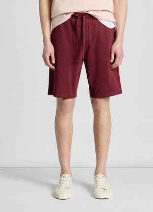 Personne portant un short bourgogne mi-long avec cordon, des baskets beige clair et un haut rose clair, debout devant un fond uni.