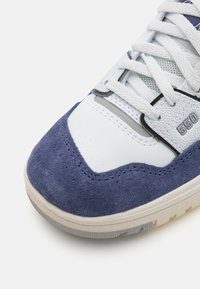 O sapato possui um верх em couro branco com detalhes em camurça azul, malha texturizada e detalhes em cinza. Língua acolchoada e fecho com atacadores. Número do modelo 550.