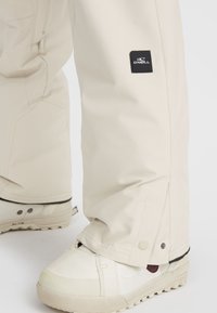 Beige snow pants feature a fabric tag with "O'Neill" logo, adjustable ankle closure with snap detail, and a relaxed fit over snow boots.