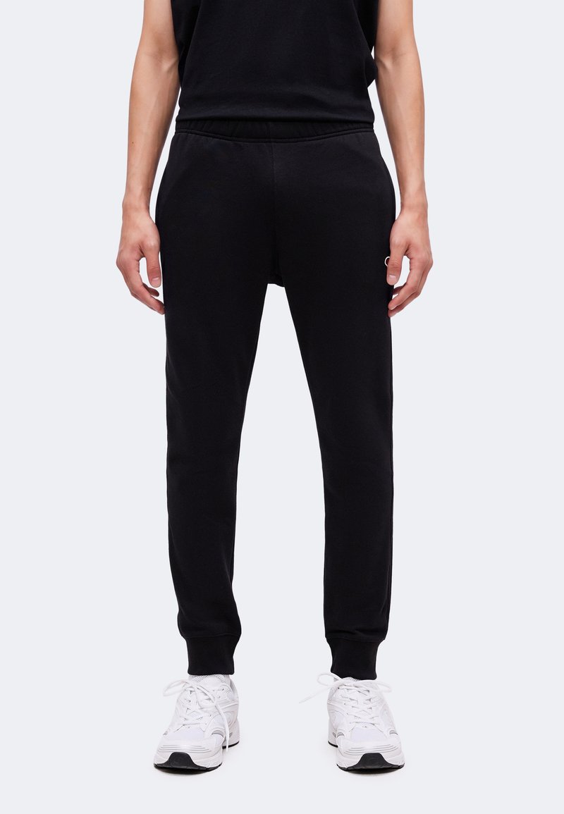 Personne portant un pantalon de jogging noir et des baskets blanches, debout devant un fond uni, les mains détendues sur les côtés.