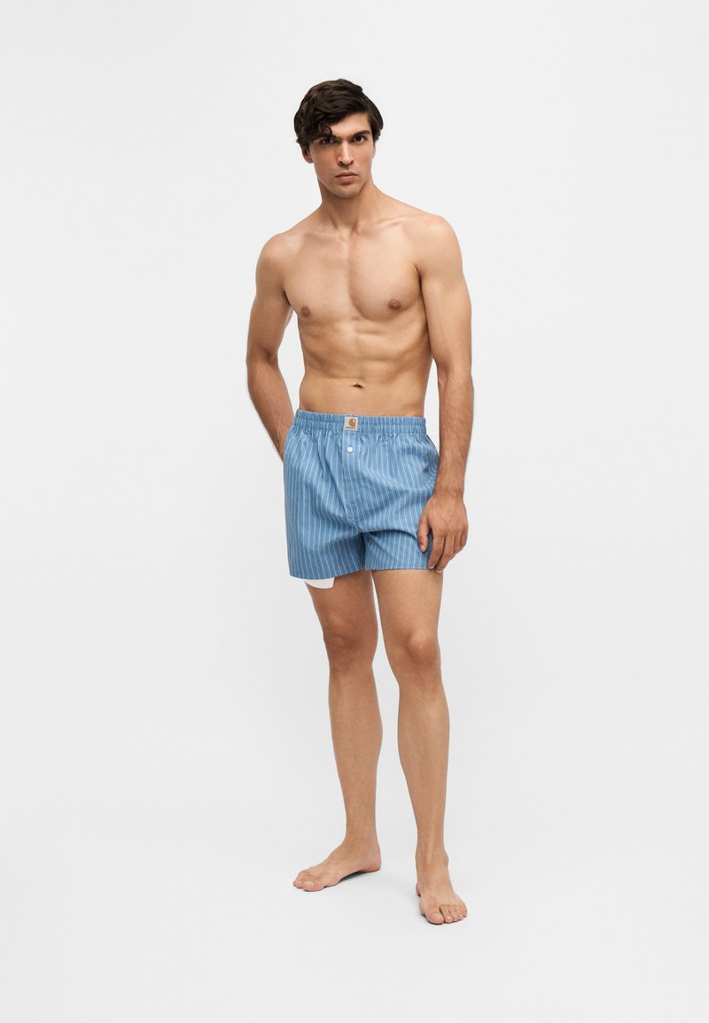 Hemdloser Mann mit dunklen Haaren, der blaue und weiße gestreifte Boxershorts trägt, barfuß vor einem schlichten hellen Hintergrund steht.