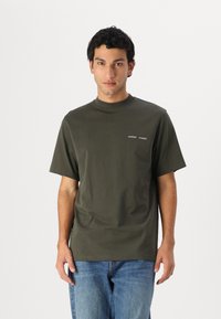 Bărbat îmbrăcat cu un tricou verde măsliniu cu mânecă scurtă, având inscripția mică albă „SAMSOE SAMSOE” și blugi albaștri, stând în fața unui fundal alb simplu.