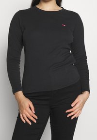 Schwarzes Langarm-T-Shirt aus Baumwolle mit Rundhalsausschnitt und kleinem roten Logo auf der linken Brust. Glatte Textur, taillierte Form.