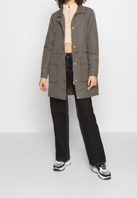 Lång grå kappa med två framfickor, knappar och dragsko i midjan, i kombination med svarta vida byxor och sneakers. Slät tygtextur.