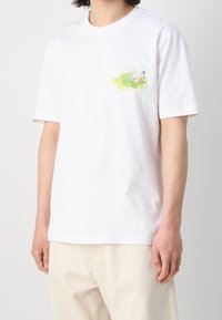 Valkoinen puuvillainen T-paita, jossa on värikäs painatus nurmikosta ja kukasta vasemmassa rinnassa. Lyhyet hihat ja rento leikkaus.