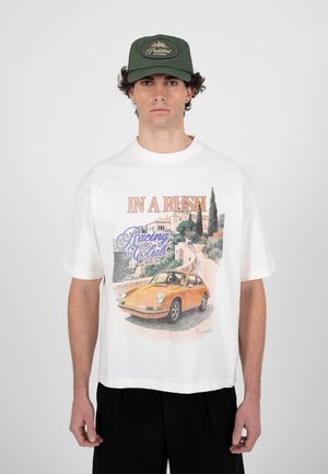 Mand iført en hvid T-shirt med et vintage gult bilmotiv og teksten "In A Rush Racing Club Berlin", kombineret med en grøn kasket og sorte bukser.