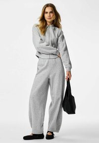 Sweat-shirt gris avec fermeture éclair à l'avant et col, associé à un pantalon large assorti, complété par des chaussures plates noires et un sac noir.