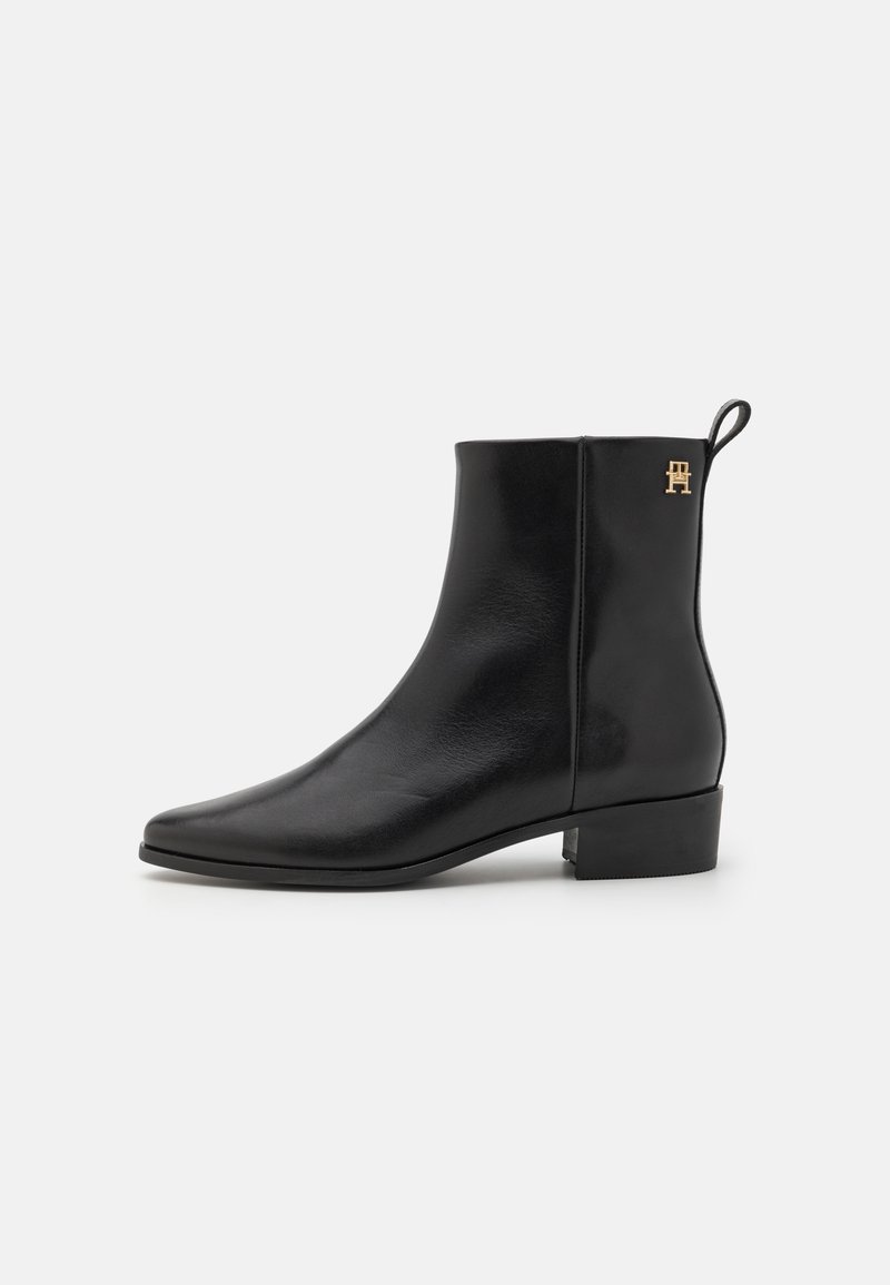 Bottine noire en cuir avec petit talon bloc, bout pointu, languette au dos et petit logo doré près de la cheville sur une surface lisse.