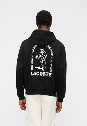 Persona vistiendo una sudadera con capucha negra Lacoste con un gráfico de tenista blanco y el texto "La leyenda de Fred, la elegancia continúa" en la parte trasera.
