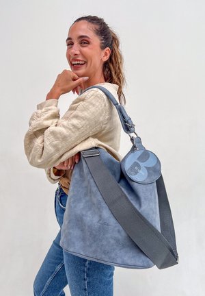 Glunderende vrouw met een paardenstaart, gekleed in een crèmekleurige jas en blauwe jeans, die een grote zachte blauwe schoudertas met een ronde label vasthoudt.