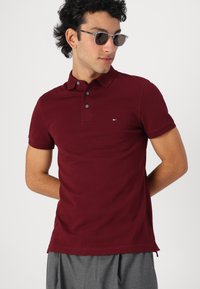 Burgundská polo košile s dvouknoflíkovým zapínáním, krátkými rukávy, malým vyšitým logem a bočními rozparky. Nosená s šedými kalhotami.