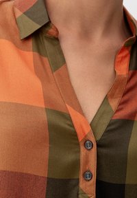 Camicia con bottoni in fantasia a quadri arancione, verde e marrone; presenta un colletto e due bottoni scuri. Tessuto liscio e intrecciato.