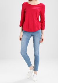 Top rojo de manga tres cuartos con un escote amplio, combinado con jeans ajustados de color azul claro. El modelo lleva zapatillas blancas. Diseño sencillo, sin estampados.