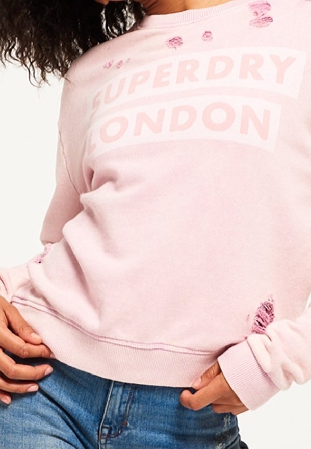 Femme portant un sweat-shirt rose clair usé avec le texte "Superdry London" et un jean bleu, posant avec les mains près des hanches.