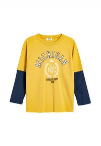 Keltainen puuvillainen pitkähihainen T-paita, jossa on laivastonsiniset hihat. Edessä on valkoinen grafiikka, jossa lukee "MICHIGAN NYC ATHLETIC DEPT 1978" sekä laakerinkukkasuunnittelu.