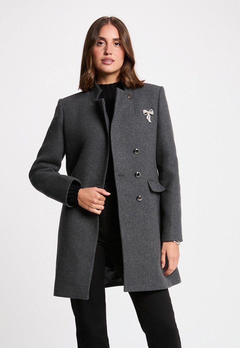 Grauer Wollmantel mit tailliertem Schnitt, hohem Kragen, sechs Knöpfen, einer Seitentasche und einem dekorativen Schleifenakzent auf der Brust.
