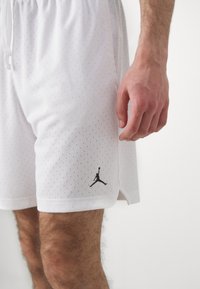 Fehér sportnadrág perforált anyagból, elasztikus derékbőséggel és fekete Jumpman logóval a bal alsó részen.