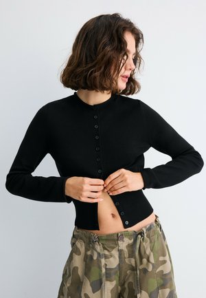 Donna con capelli ricci fino alle spalle che si sta abbottonando un cardigan nero a maniche lunghe, indossa pantaloni larghi mimetici su uno sfondo semplice.