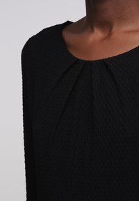 Top de tela negra texturizada con escote redondo y detalle sutil de pliegues. El patrón de la tela añade profundidad al diseño general.