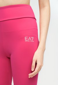 Κοντινή λήψη του κάτω μέρους του κορμού και του δεξιού χεριού ενός ατόμου που φοράει φωτεινά ροζ κολάν με λευκό λογότυπο EA7 Emporio Armani στο ισχίο.