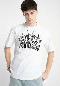 Biała koszulka z krótkim rękawem z czarną grafiką przedstawiającą trzy głowy psów, nad słowem "BEZLITOSNY" w pogrubionym, stylizowanym kroju pisma.