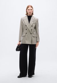 Διπλόσαρκο σακάκι με μοτίβο houndstooth σε γκρι και μαύρο, συνδυασμένο με μαύρο παντελόνι με φαρδιά μπατζάκια· το μοντέλο κρατά μια μικρή μαύρη τσάντα.