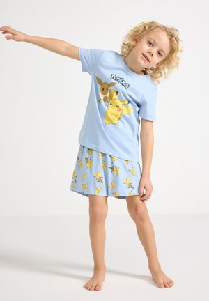 Barn iført lyseblå Pokémon-trøje og matchende nederdel, står barfodet med den ene arm strakt ud til siden mod en ensfarvet baggrund.