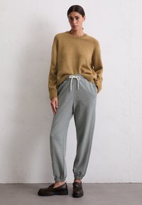 Khaki gebreide trui gecombineerd met lichte grijze tapered joggers met een trekkoord en elastische cuffs, gedragen met donkerbruine instappers.