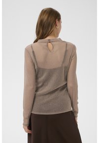 Permatomas, ilgomis rankovėmis megztinis iš blizgaus šviesiai rudos spalvos audinio, su raktelio formos iškirpte nugaroje ir aptempiančia forma, dėvimas ant tamsesnio drabužio.