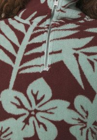 Gros plan sur un tissu polaire avec de grands motifs floraux et feuillus en bleu clair et marron, avec une fermeture éclair verticale argentée.