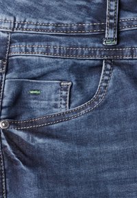 Blauwe spijkerbroek met een voorzak, voorzien van contrasterende stiksels en een klein groen accent. De stof heeft een gladde textuur met een lichte glans.