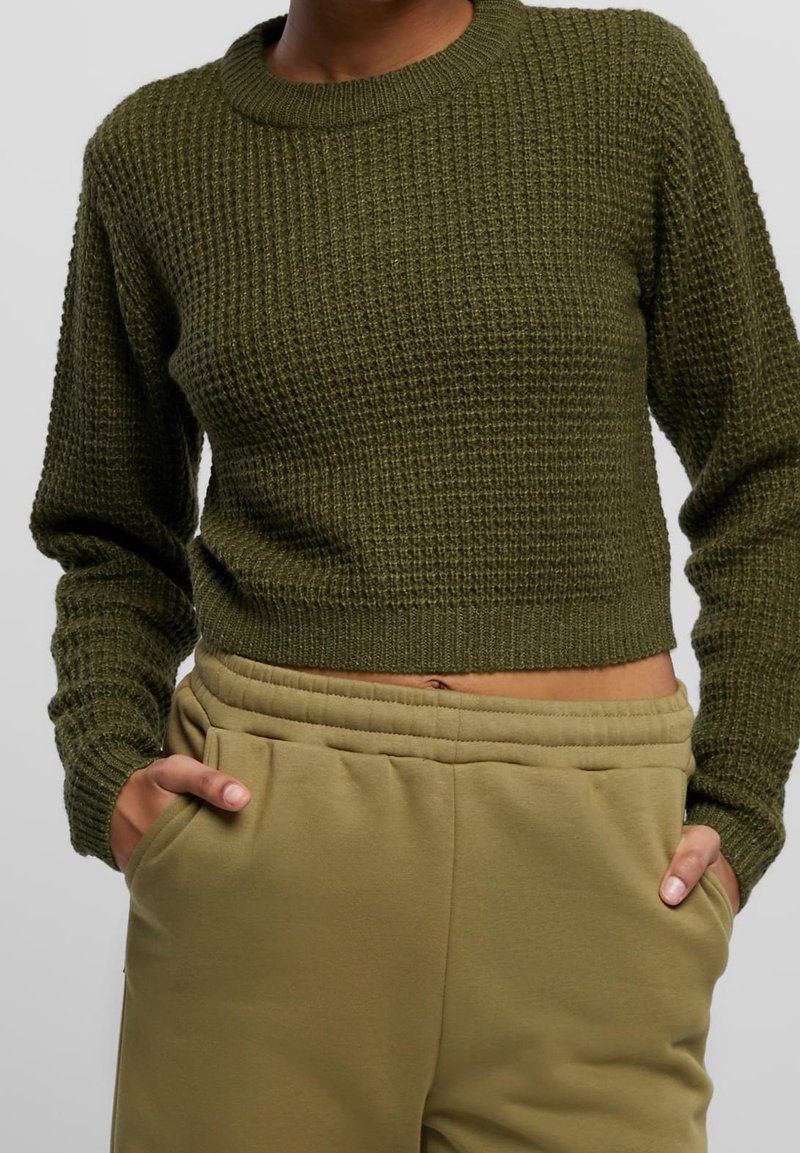 Pull court en maille texturée vert olive porté avec un pantalon de jogging beige taille haute, mains dans les poches, sur fond gris uni.